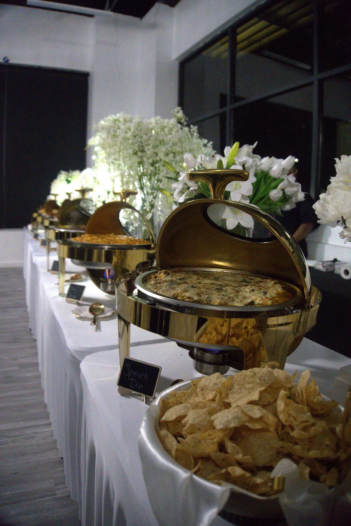A long buffet of gold chafing dishes and white florals set for a wedding reception