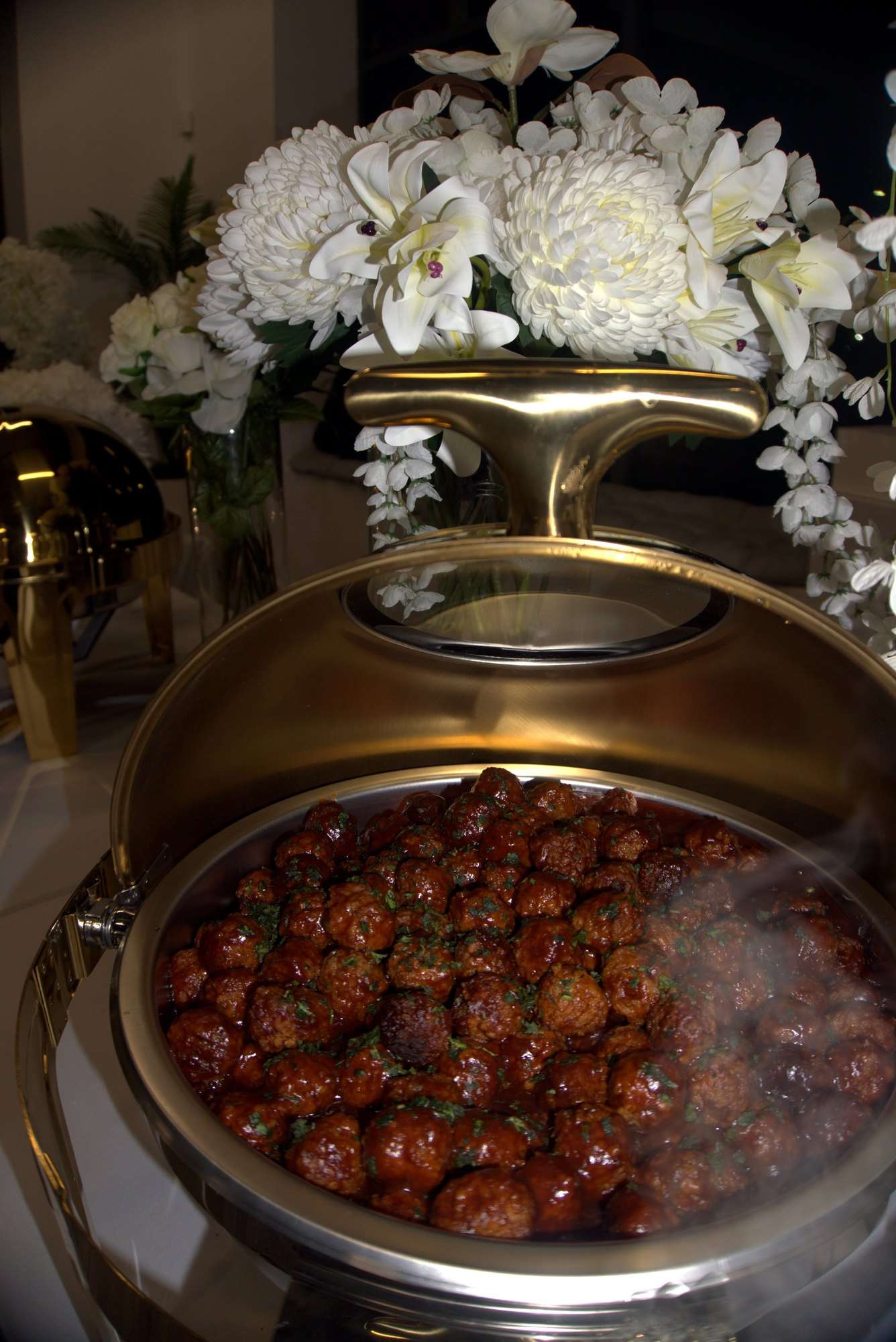 Glazed meatballs steaming in a gold chafing dish with white flowers behind