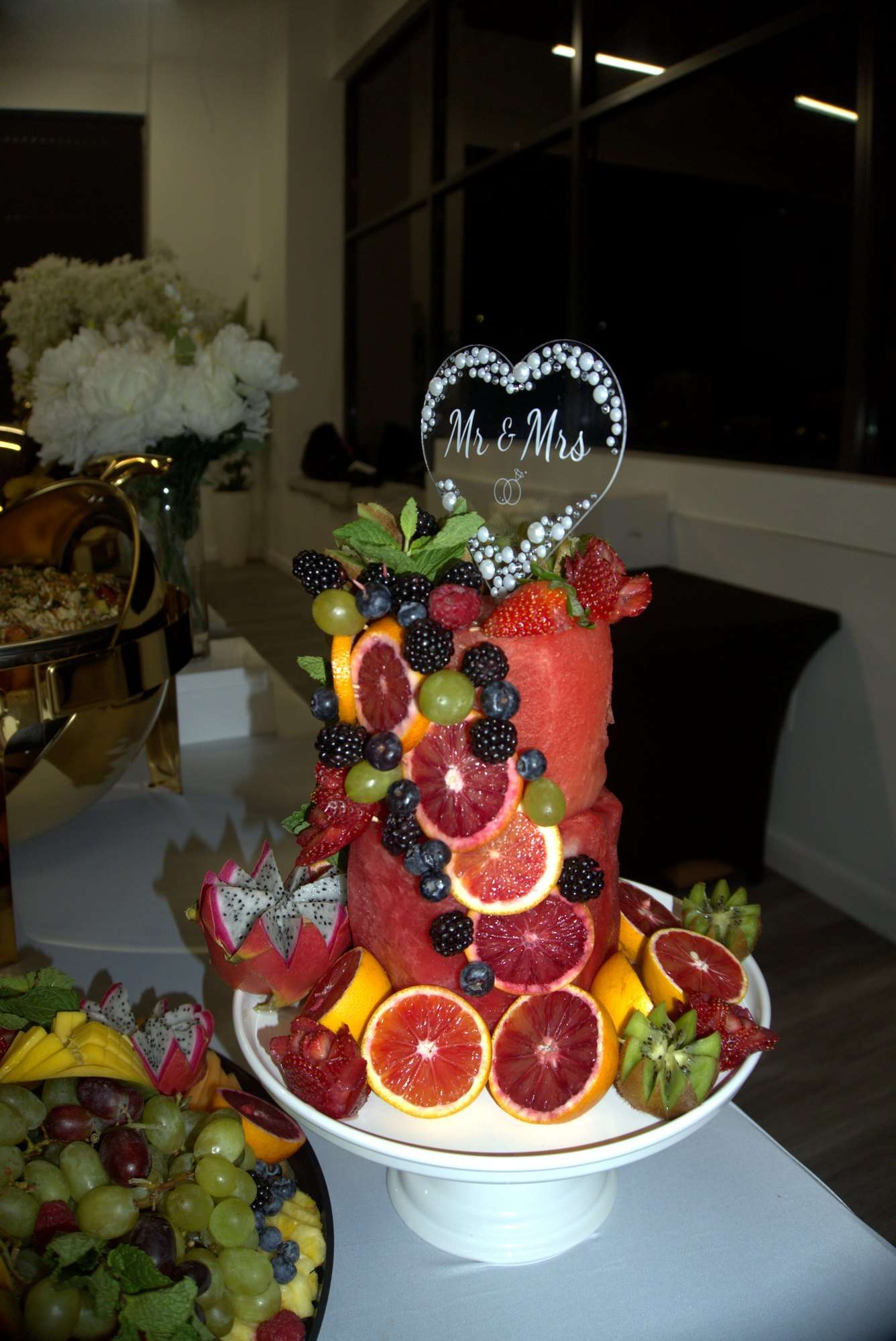 A watermelon tower decorated with blood oranges and berries under a Mr & Mrs topper