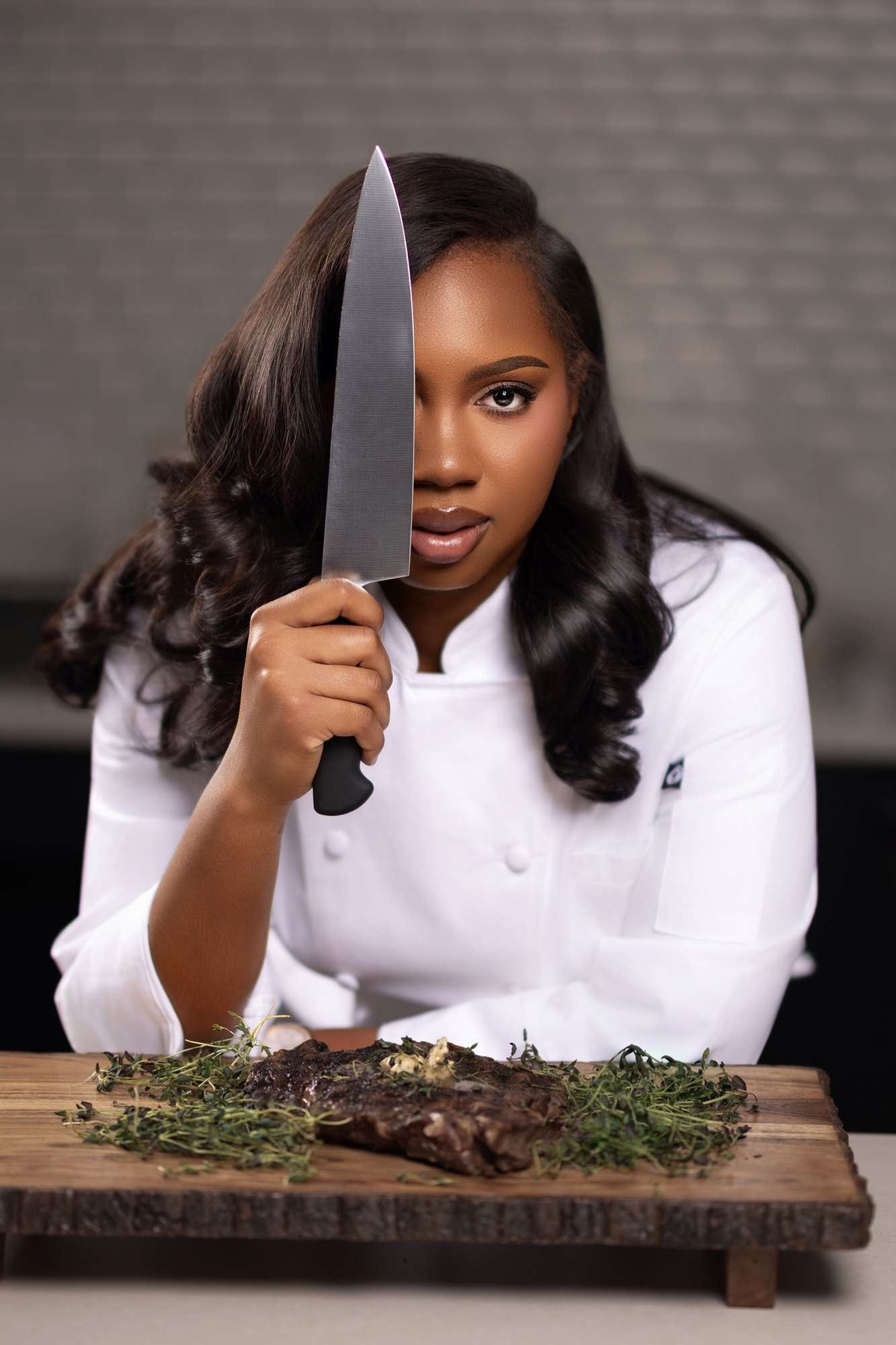 Jordan Ali-Taylor in chef whites holding a chef’s knife upright beside a steak on a wooden board