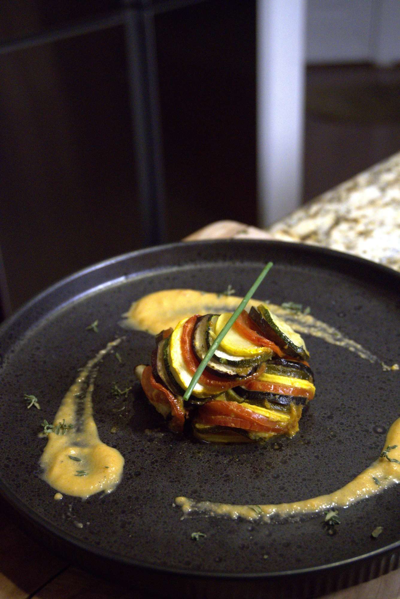 A tower of layered ratatouille with a single chive, swept with a golden sauce on a black plate