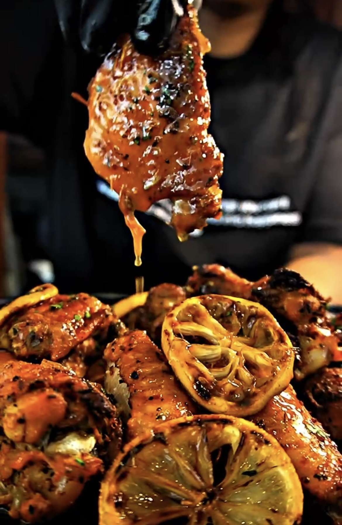 A glazed chicken wing lifted from a platter of wings with charred lemon, glaze dripping